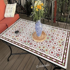 May include: A rectangular outdoor coffee table with a mosaic tile top featuring a geometric pattern in red, green, and white. The table has a black metal frame and is set on a wooden deck. A vase with flowers sits on the table.