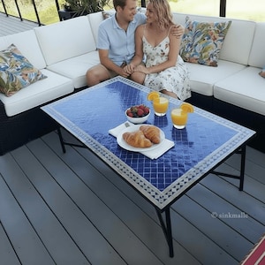 May include: A rectangular outdoor coffee table with a blue tile top and a mosaic border. The table has black metal legs and is set with breakfast items, including croissants, fruit, and orange juice. A couple is seated on a white sofa in the background.