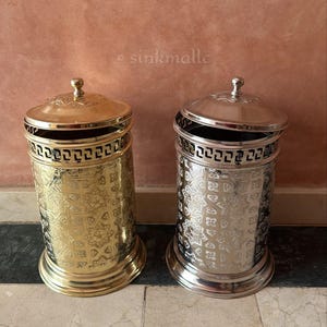 May include: Two cylindrical waste bins with lids, one in a polished gold finish and one in a polished silver finish. Both feature an intricate embossed geometric pattern on the body and a Greek key border near the top rim. Each lid has a small knob.