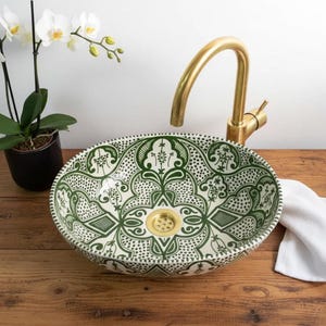 May include: An oval ceramic vessel sink featuring a green and white intricate geometric pattern. The sink includes a gold-toned metal drain and is paired with a matching gold-toned curved faucet. The design covers the interior basin surface.