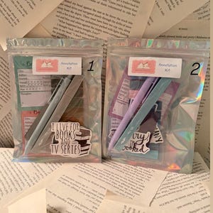 Puede incluir: Dos bolsas de plástico transparente que contienen kits de anotación, cada una con bolígrafos, pegatinas y tarjetas. Las bolsas están sobre un fondo de páginas de libros. Una bolsa tiene el texto "I LIVE FOR BOOKS AND TV SERIES".