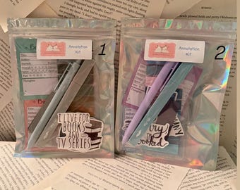 Peut inclure: Deux sacs en plastique transparents contenant des kits d'annotation, chacun avec des stylos, des autocollants et des cartes. Les sacs sont posés sur un fond de pages de livres. Un sac porte l'inscription "I LIVE FOR BOOKS AND TV SERIES".