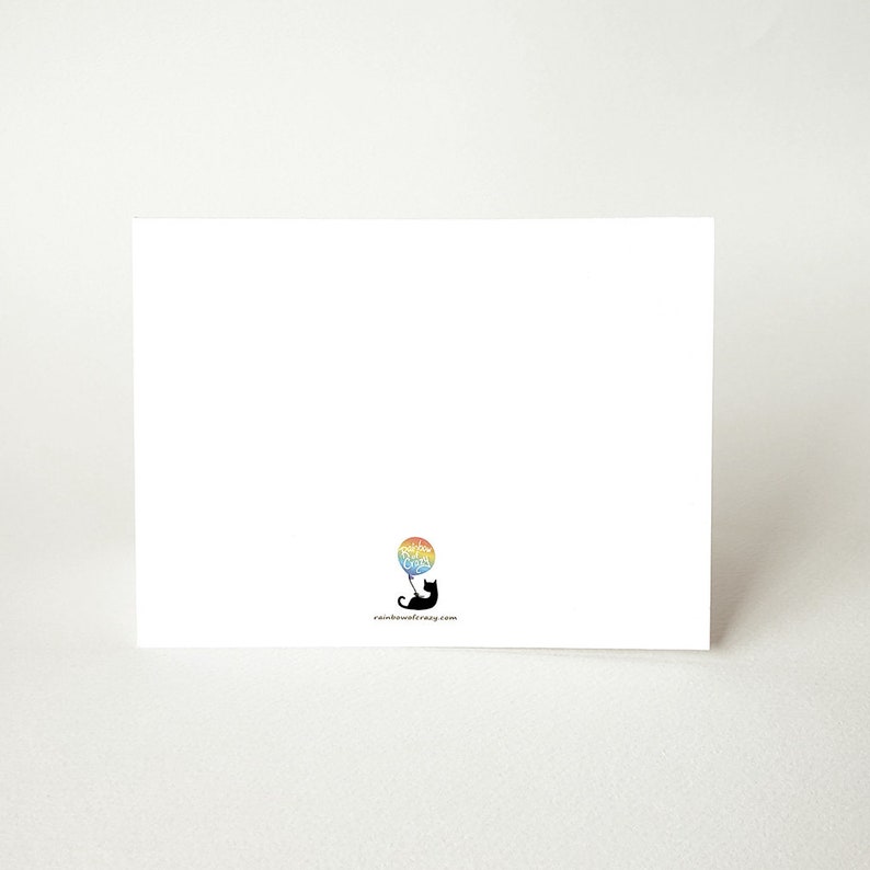 May include: A blank white card with a black silhouette of a cat holding a rainbow balloon. The text "rainbowcatmag.com" is printed in black at the bottom of the card.