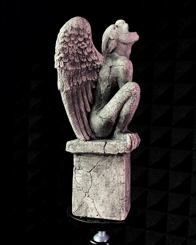 K&ouml;nnte beinhalten: Graue Gargoyle-Statue mit gro&szlig;en, detaillierten Fl&uuml;geln, die auf einem verwitterten Sockel sitzt. Die Statue befindet sich in einer hockenden Position, wobei sichtbare Risse zu ihrem gealterten Aussehen beitragen. Der Hintergrund ist schwarz.