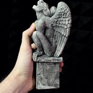 K&ouml;nnte beinhalten: Graue Gargoyle-Statue mit detaillierten Fl&uuml;geln, auf einem Steinsockel. Die Statue hat ein verwittertes Aussehen mit sichtbaren Rissen und einer strukturierten Oberfl&auml;che. Die Gargoyle befindet sich in einer hockenden Position mit offenem Mund.