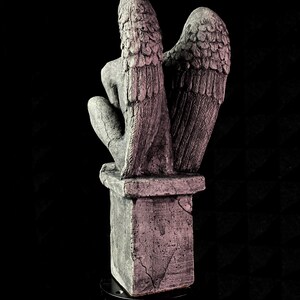 K&ouml;nnte beinhalten: Graue Steinfigur eines Engels mit gro&szlig;en, detaillierten Fl&uuml;geln. Der Engel kniet auf einem quadratischen Sockel. Die Statue hat ein verwittertes Aussehen mit sichtbaren Rissen und Texturen, vor schwarzem Hintergrund.