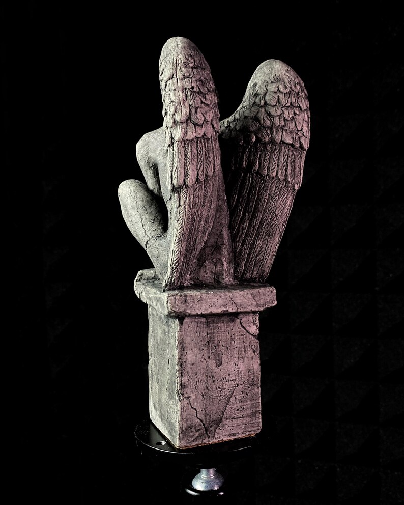K&ouml;nnte beinhalten: Graue Steinfigur eines Engels mit gro&szlig;en, detaillierten Fl&uuml;geln. Der Engel kniet auf einem quadratischen Sockel. Die Statue hat ein verwittertes Aussehen mit sichtbaren Rissen und Texturen, vor schwarzem Hintergrund.