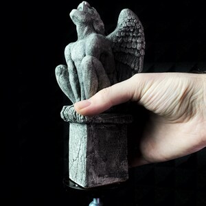 K&ouml;nnte beinhalten: Eine graue Gargoyle-Statue mit detaillierten Fl&uuml;geln und einer sitzenden Pose. Die Statue steht auf einem quadratischen Sockel mit einem strukturierten, verwitterten Aussehen. Die Gargoyle besteht aus einem steinartigen Material und ist vor einem schwarzen Hintergrund platziert.