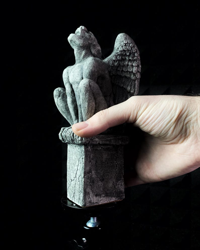 K&ouml;nnte beinhalten: Eine graue Gargoyle-Statue mit detaillierten Fl&uuml;geln und einer sitzenden Pose. Die Statue steht auf einem quadratischen Sockel mit einem strukturierten, verwitterten Aussehen. Die Gargoyle besteht aus einem steinartigen Material und ist vor einem schwarzen Hintergrund platziert.