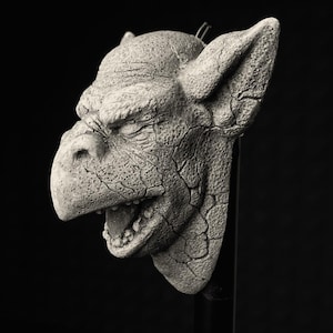 Puede incluir: Escultura en escala de grises de una cabeza de gárgola con texturas detalladas y boca abierta. El material, similar a la piedra, presenta grietas e imperfecciones, con orejas prominentes y una expresión severa. La escultura está montada sobre un soporte negro.
