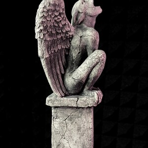 K&ouml;nnte beinhalten: Graue Gargoyle-Statue mit gro&szlig;en Federfl&uuml;geln, die auf einem quadratischen Sockel sitzt. Die Statue hat ein verwittertes Aussehen mit sichtbaren Rissen, vor einem schwarzen Hintergrund. Die Gargoyle sitzt und blickt nach oben.