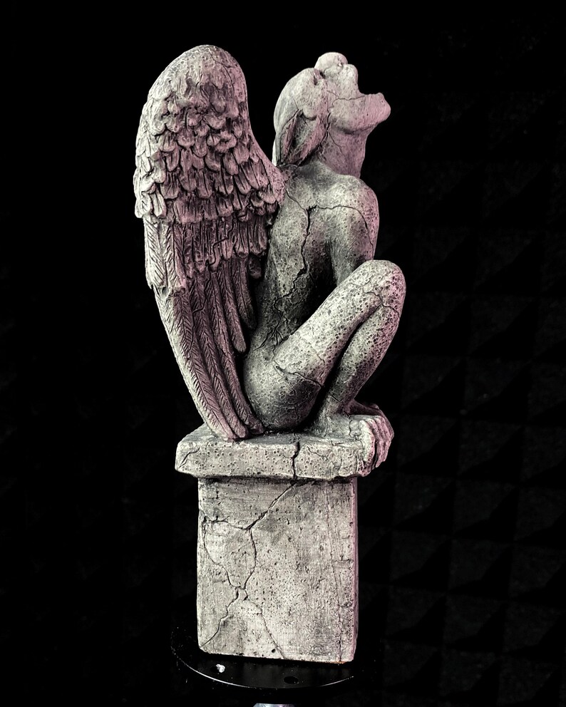 K&ouml;nnte beinhalten: Graue Gargoyle-Statue mit gro&szlig;en Federfl&uuml;geln, die auf einem quadratischen Sockel sitzt. Die Statue hat ein verwittertes Aussehen mit sichtbaren Rissen, vor einem schwarzen Hintergrund. Die Gargoyle sitzt und blickt nach oben.