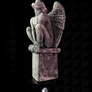 K&ouml;nnte beinhalten: Eine graue Gargoyle-Statue mit gro&szlig;en Fl&uuml;geln, die auf einem quadratischen Sockel sitzt. Die Statue hat strukturierte Oberfl&auml;chen und Risse und ist vor einem dunklen Hintergrund platziert. Die Gargoyle blickt nach oben.