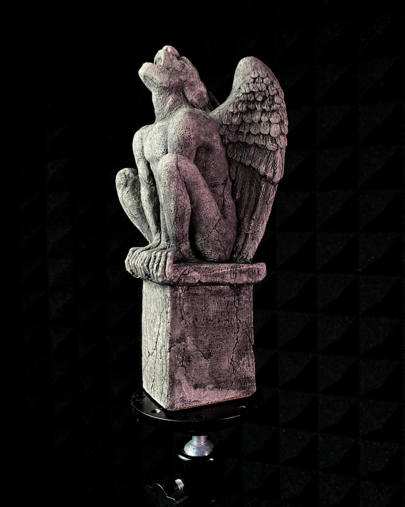 K&ouml;nnte beinhalten: Eine graue Gargoyle-Statue mit gro&szlig;en Fl&uuml;geln, die auf einem quadratischen Sockel sitzt. Die Statue hat strukturierte Oberfl&auml;chen und Risse und ist vor einem dunklen Hintergrund platziert. Die Gargoyle blickt nach oben.