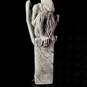 Puede incluir: Estatua gris y desgastada de una criatura con una gran cabeza, alas y tentáculos. La estatua está posada sobre una base rectangular agrietada. La escultura tiene una superficie detallada y texturizada sobre un fondo negro.