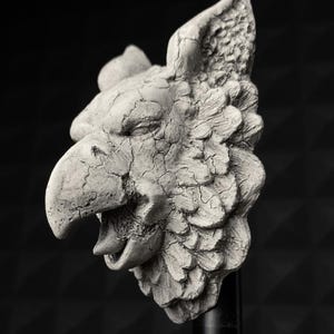 Puede incluir: Escultura en escala de grises de la cabeza de un grifo, con pico de ave y detalles de plumas. La escultura tiene una textura agrietada y envejecida, montada sobre una varilla negra sobre un fondo oscuro. La boca del grifo está abierta.