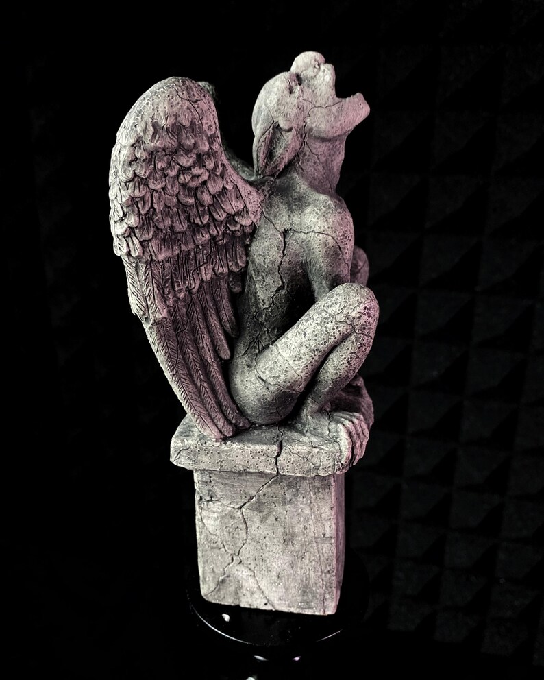 K&ouml;nnte beinhalten: Graue Gargoyle-Statue mit gro&szlig;en, detaillierten Fl&uuml;geln und einem rissigen, verwitterten Aussehen. Die Statue sitzt auf einem quadratischen Sockel, mit zur&uuml;ckgeneigtem Kopf und offenem Mund. Die Skulptur ist aus Stein.