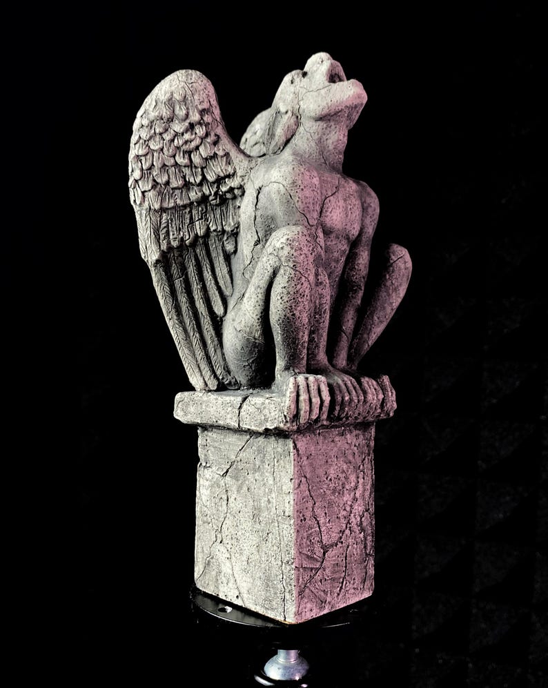 K&ouml;nnte beinhalten: Graue Gargoyle-Statue mit gro&szlig;en, detaillierten Fl&uuml;geln und rissigem, verwittertem Aussehen. Die Statue thront auf einem quadratischen Sockel mit &auml;hnlicher Textur, vor schwarzem Hintergrund. Die Gargoyle blickt nach oben.