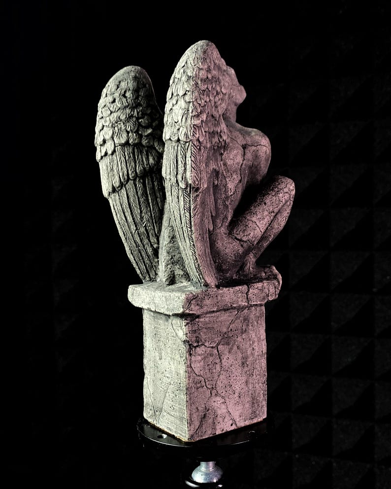 K&ouml;nnte beinhalten: Steinstatue einer gefl&uuml;gelten Figur, m&ouml;glicherweise eines Engels oder einer Wasserspeiers, in kniender Position. Die Fl&uuml;gel sind detailliert ausgearbeitet. Die Statue ist grau mit rosa Farbt&ouml;nen, der Sockel weist Risse auf.