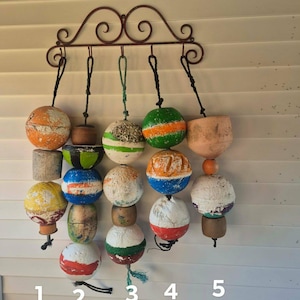 May include: A collection of weathered, colorful buoys hanging from a decorative wrought iron wall mount. The buoys are painted in various colors, including orange, green, blue, and white, and are suspended by ropes. A rustic nautical theme.