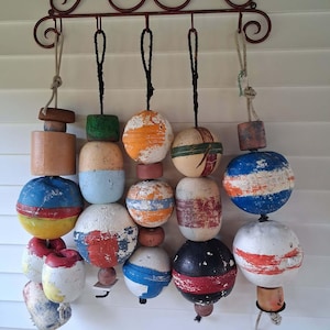 May include: A decorative wall-mounted metal rack with scrollwork details, featuring five hanging ropes. Each rope holds a vertical arrangement of weathered, multi-colored foam fishing buoys in various shapes, including spheres and cylinders.