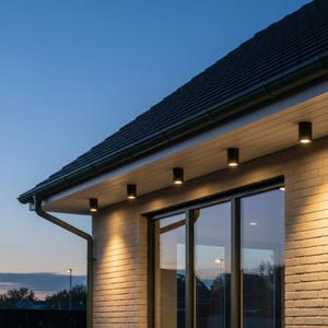 Puede incluir: Vista exterior de una casa al anochecer. La imagen muestra una sección del tejado, una pared de ladrillo y una gran ventana. Varias luces cilíndricas negras están montadas bajo el alero del tejado, iluminando la pared.