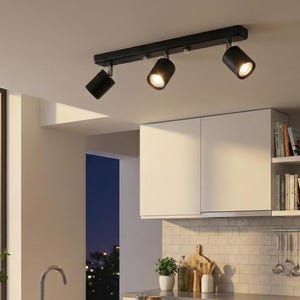 May include: A black, three-light track lighting fixture. Each light is cylindrical and adjustable, with a bright, focused beam. The fixture is mounted on a white ceiling, above a kitchen setting. The lights are turned on.