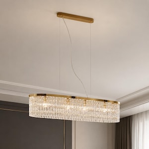 Puede incluir: Una lámpara colgante de forma ovalada con un marco de metal dorado y varias filas de cristales de vidrio transparentes y rectangulares. La lámpara está suspendida del techo mediante cables finos unidos a un dosel rectangular dorado.