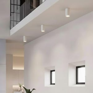 Puede incluir: Luces de techo cilíndricas blancas instaladas en un techo blanco. Las luces están espaciadas uniformemente e iluminan un interior moderno con paredes blancas y pequeñas ventanas cuadradas. La imagen muestra un diseño minimalista.