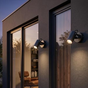 Pode incluir: Dois holofotes pretos para exteriores montados numa parede cinza escuro. Cada holofote tem uma cabeça cilíndrica e uma base circular. As luzes estão anguladas para baixo, iluminando a parede abaixo. A cena é ao ar livre, com um vislumbre de uma sala visível através de uma janela.