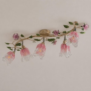 Può includere: Una lampada da soffitto con una struttura metallica curva a forma di ramo con finitura anticata. È decorata con piccole foglie verdi e delicati accenti di rose viola. Pendono cinque paralumi in vetro a forma di fiori rosa e bianchi.
