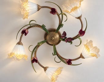 Op de afbeelding: Een plafondlamp met vijf lichtpunten, voorzien van gebogen bronskleurige metalen armen versierd met groene bladeren en kleine donkerrode bloemknoppen. De lampen hebben de vorm van witte en amberkleurige glazen bloemen.