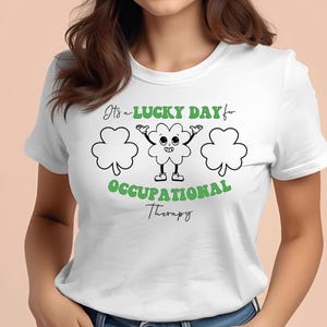 以下が含まれることがあります： 「It's a LUCKY DAY for OCCUPATIONAL Therapy」の文字が緑色でプリントされた白いTシャツ。デザインには、顔と腕が付いたものを含む3つの緑色のシャムロックが含まれています。Tシャツは人が着ています。