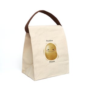 May include: A beige canvas lunch bag with a brown handle. The bag features a smiling cartoon potato with the words "Positive Potato" printed above and below the image. The bag is designed to carry food and drinks.