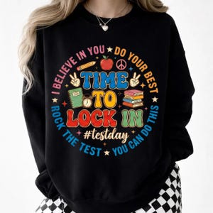 Puede incluir: Sudadera negra con un gráfico colorido que dice "I believe in you, do your best, time to lock in, #testday." El diseño incluye una manzana, libros, un lápiz y un signo de la paz. La sudadera tiene cuello redondo.