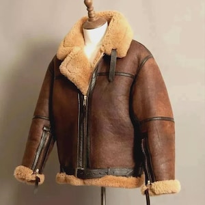 Puede incluir: Chaqueta de cuero marrón con cuello y forro de borrego. La chaqueta tiene cierre de cremallera, cinturón en la cintura y puños con cremallera. La chaqueta se exhibe en un maniquí.