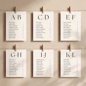 May include: Six off-white rectangular seating chart cards hanging on two horizontal strings with wooden clips. Each card features bold black alphabetical headers from A-L and lists names with corresponding table numbers in a clean serif font.