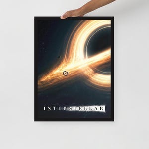 Può includere: Un poster incorniciato con una rappresentazione di un buco nero con luce arancione e gialla vorticosa. La parola "INTERSTELLAR" è stampata in bianco nella parte inferiore dell'immagine. Il poster è tenuto da una persona.