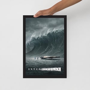 May include: A framed art print featuring a dramatic ocean scene with a massive wave and two figures in the water. The print has the word "INTERSTELLAR" at the bottom. The artwork is held by a person's hand.
