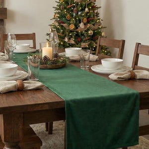May include: A festive dining table set for a holiday meal. The dark wooden table is adorned with white dishes, linen napkins, and a green table runner. A lit candle and a decorated Christmas tree are in the background.