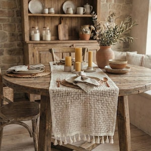 May include: A dining table set for a meal with a rustic aesthetic. The round wooden table is set with plates, napkins, and candles. A woven runner with tassels runs down the centre. A wooden hutch in the background displays dishes and jars.