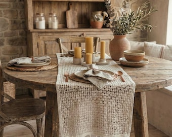 Può includere: Un tavolo da pranzo rustico apparecchiato per un pasto. Il tavolo rotondo in legno è apparecchiato con piatti, tovaglioli e candele. Un runner intrecciato con nappe corre al centro. Una credenza in legno sullo sfondo espone piatti e barattoli.