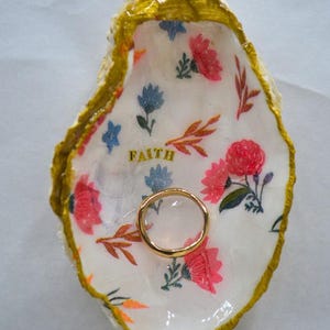 May include: A decorative oyster shell ring dish with a gold rim and floral pattern. The interior features pink and blue flowers, with the word "Faith" in gold lettering. A gold ring rests inside the dish.