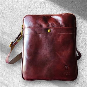 Può includere: Borsa a tracolla in pelle bordeaux con tasca frontale e chiusura con bottone dorato. La borsa ha una tracolla fantasia e un piccolo logo nell'angolo in basso a destra. La borsa è rettangolare.