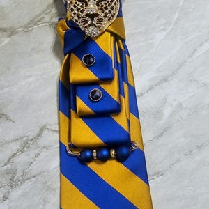 Peut inclure: Cravate rayée bleue et or ornée d'une broche tête de léopard décorative. La broche est dorée avec des accents noirs et des strass transparents. La cravate comporte également une épingle perlée bleue et deux boutons bleus.