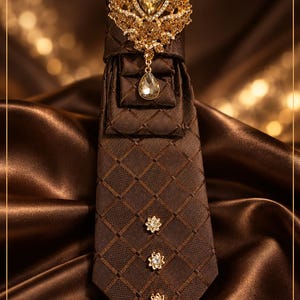 May include: A dark brown necktie with a grid pattern, featuring three small crystal flower accents and a large gold-toned brooch with a teardrop crystal. The text reads L Snell Noire Collection, Espresso Empress, and Rich in elegance. Crowned in confidence.