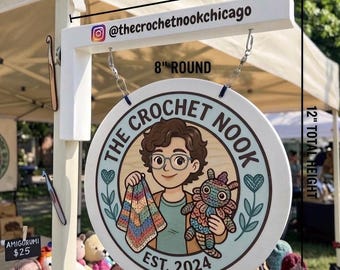 May include: A white wooden hanging sign measuring 10 inches wide and 12 inches in total height. The round sign is 8 inches in diameter and features an illustration of a person holding a colorful blanket and a creature with the text The Crochet Nook Est. 2024.