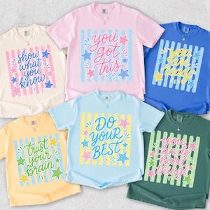 May include: A collection of six short-sleeved t-shirts in various colors, each featuring a unique inspirational message. The shirts display phrases like "Show what you know" and "You got this" in colorful, playful fonts, with star and stripe designs.