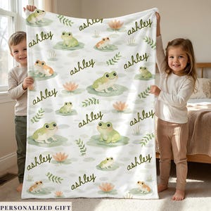 May include: A white blanket featuring a repeating pattern of green and orange frogs on lily pads, with the name "Ashley" in a handwritten font. The blanket also includes illustrations of water lilies and foliage.