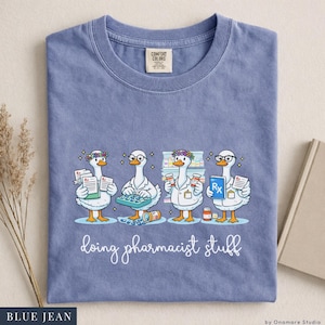 May include: Blue jean Comfort Colors t-shirt featuring a whimsical illustration of four geese dressed as pharmacists. The geese are holding prescription pads, medicine bottles, and a keyboard, with the phrase "doing pharmacist stuff" below.